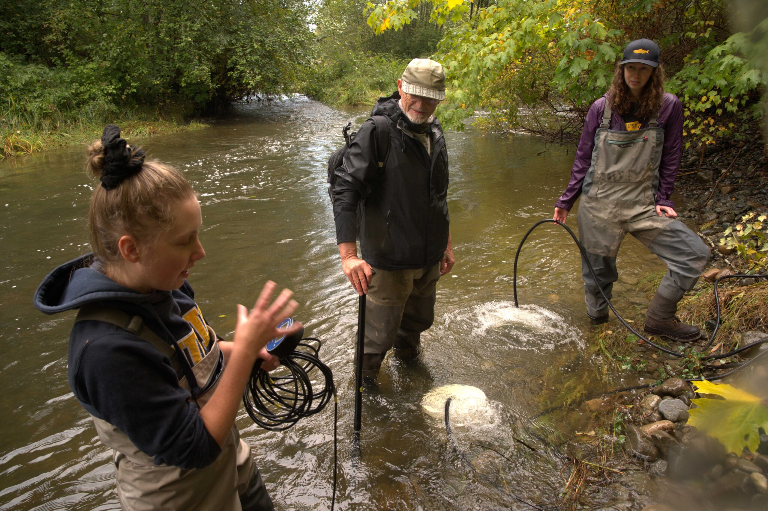Katie Gair (TRRS) describes the deployment of portable aeration units in refuge pools of the Tsolum River to increase the survival of pink salmon adults returning to spawn. Allan Chamberlain (TRRS) and Jane Pendray (PSF) stand in the Tsolum River holding tubes leading to aeration units that are actively bubbling oxygen into the water.