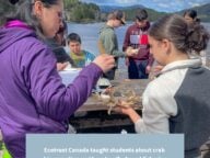 A teacher hands a crab to a high school student in an outdoor classroom at a table surrounded by students