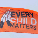 An orange flag with the text "Every Child Matters" and two black and white feathers flies against the backdrop of a blue sky