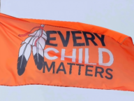 An orange flag with the text "Every Child Matters" and two black and white feathers flies against the backdrop of a blue sky