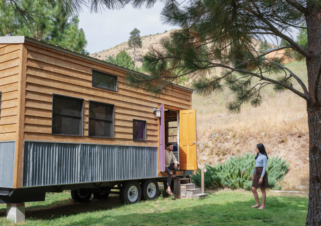 A wooden tiny home is parked on a grass expanse, where a woman stands looking at a man in the door to the RV